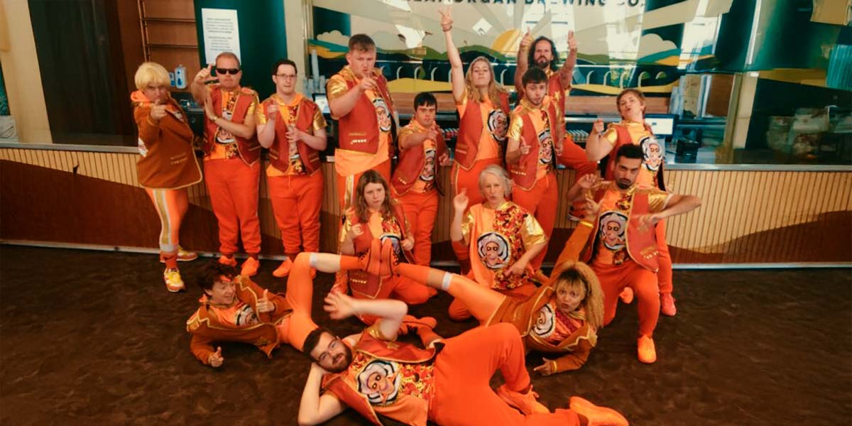 Fifteen people are dressed identically in bright orange trousers and shoes with open waistcoats over orange t-shirts bearing the print of a man wearing a Stetson and sunglasses. They pose in front of a stage, some lying on the floor, others standing and pointing to the camera.
