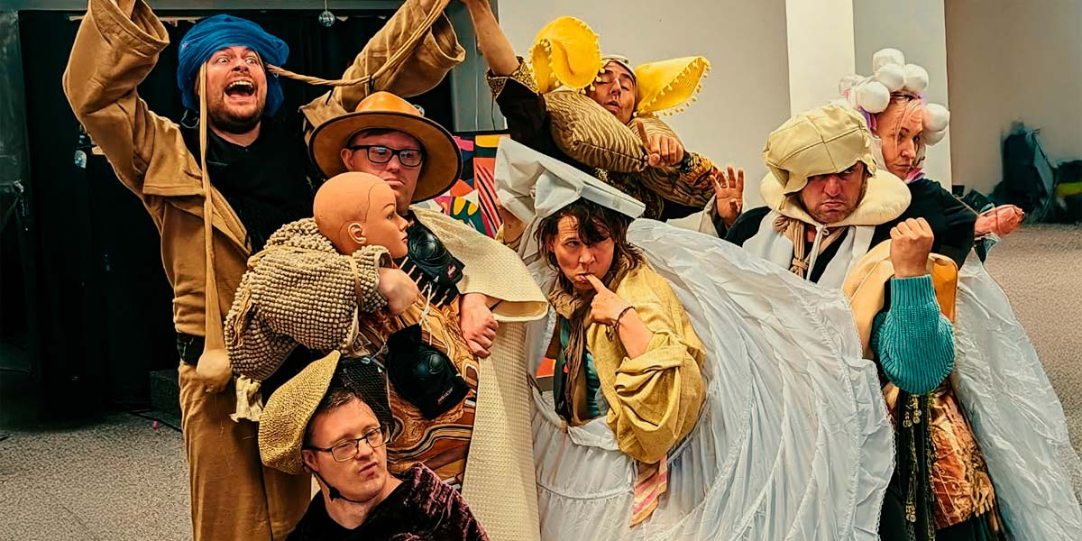 Severn members of Truth pose in thrown-together costumes of pillows, curtains and robes. They all have hats- flat caps, sun hats and home-made ones of cloth, place mats and polystyrene balls. One man grins manically and holds a large maraca, wrapped in a pair of tan tights, above his head.