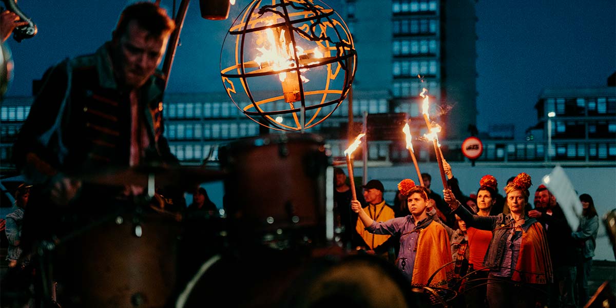 Under a night sky, a group wearing orange capes and pompoms on their heads hold up flaming torches. Their faces are sad in the soft light. In the foreground, a globe-shaped cage holding fire burns brightly, whilst a downcast drummer plays his drums.