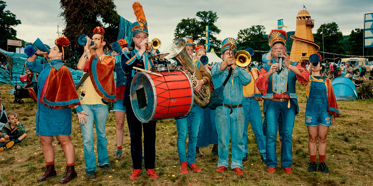 In a festival field, a group of people dressed in denim with red pompoms on their heads, hold brass instruments or blue cones to their mouths.