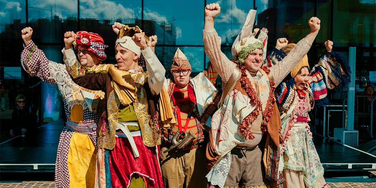 Five performers in colourful outfits made from household items such as tablecloths, curtains and bedspreads cluster together and look outwards, grinning. Their arms are raised in fists.