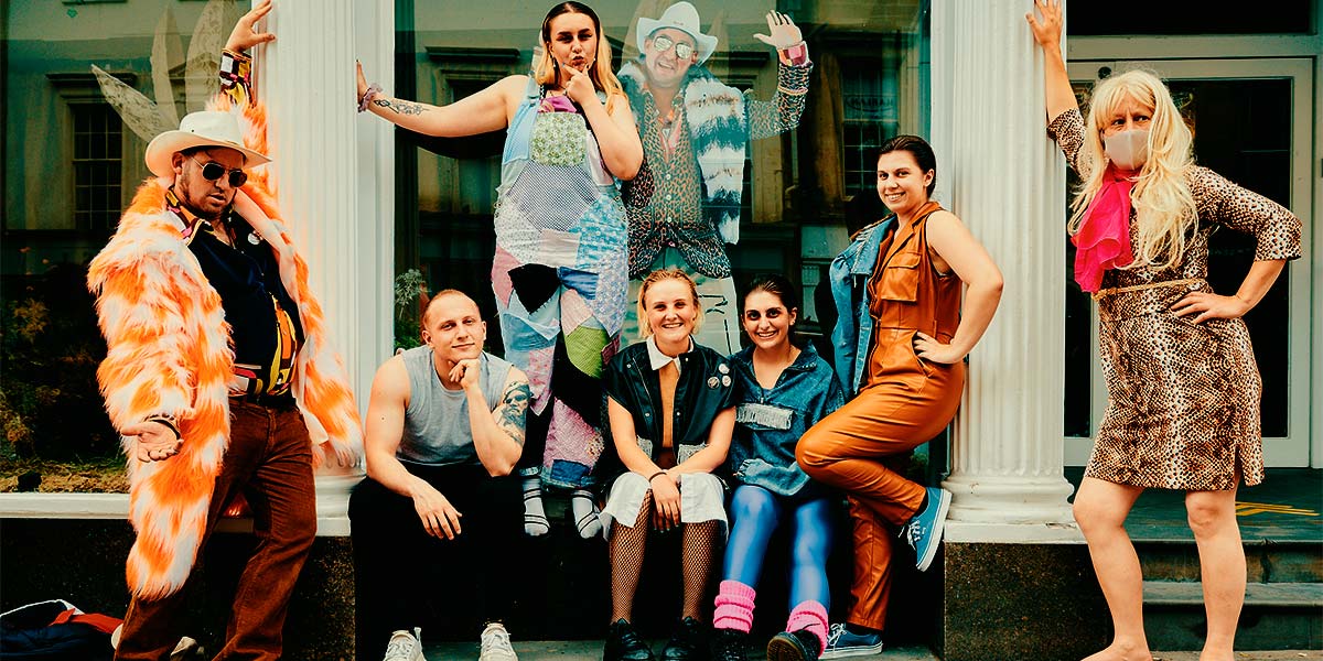 A group of smiling students sit and stand around a shop window. Posing enticingly at either side of them is a man dressed in a huge, orange-mottled fur coat, white Stetson hat and sunglasses and a woman wearing a facemask and dressed in a leopard print dress, blond wig and bare feet. Inside the shop window is the grinning cardboard replica of the man in the Stetson hat.
