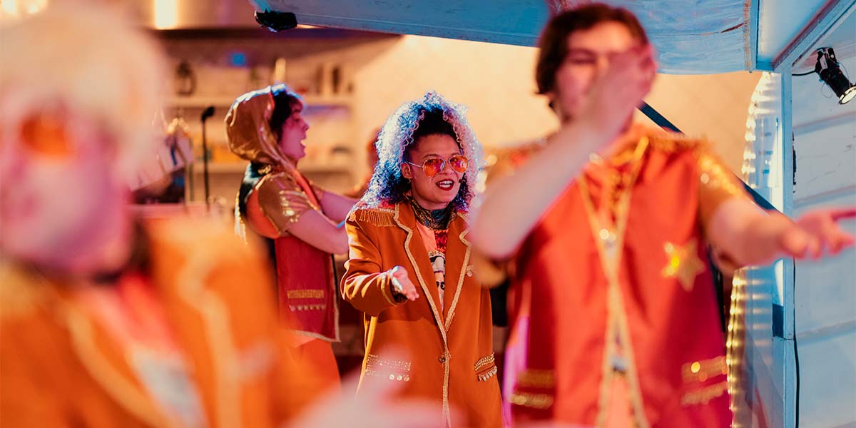  A group of people dressed in Club Supreme outfits sing and dance beside a caravan. Some are out of focus and appear as a blur of orange. Light glints off the gold embellishments on their costumes.