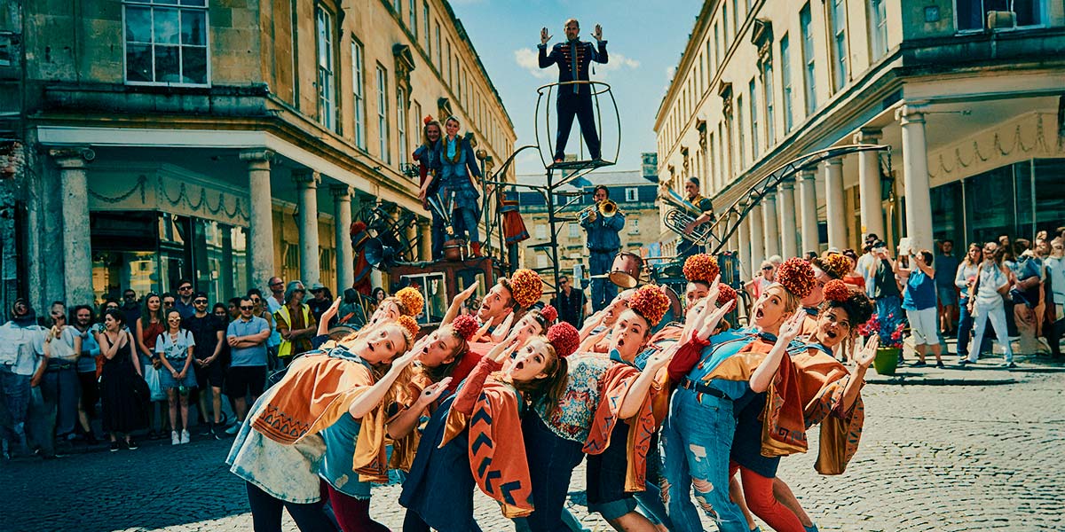 In a town square, a group of young performers lean backwards in a clump, their arms raised at right angles and their mouths open in an ‘o’. They wear orange pompoms on their heads. On an elevated platform behind them, a man in a blue velvet suit has his arms raised in a similar gesture.