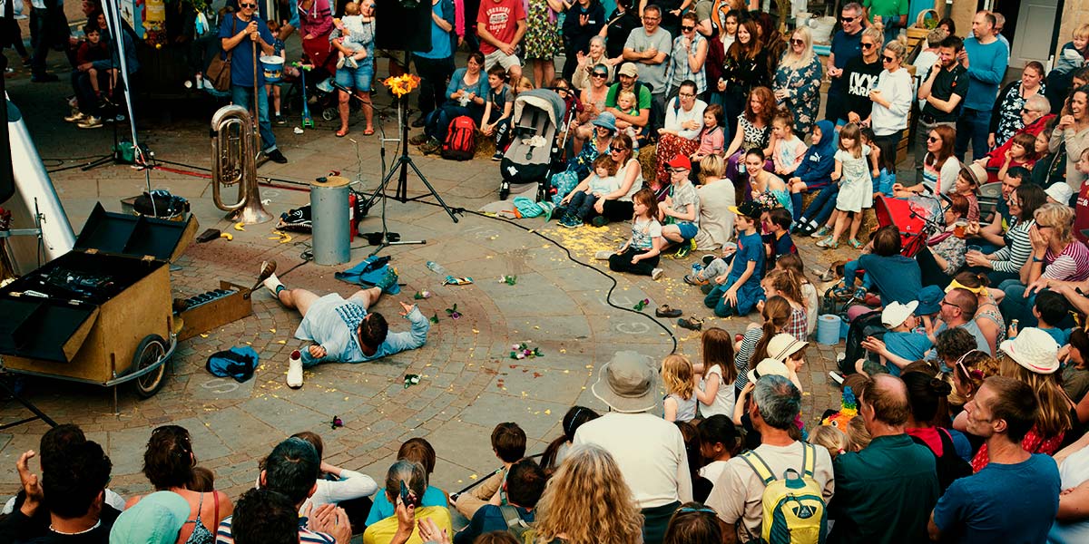 A large audience watch a man lying on his back. He has no trousers on and the floor around him is strewn with mess. A small fire burns by a speaker.