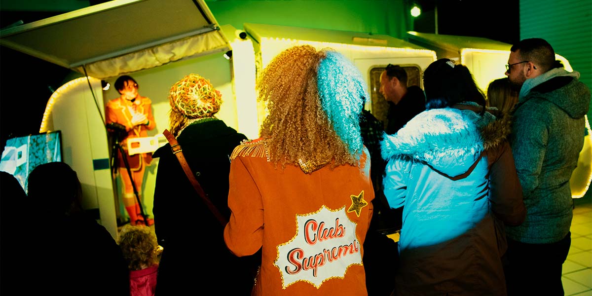 A group of people watch a Club Supreme performer standing inside a caravan. The back of another performer is in focus, with the words Club Supreme emblazoned on her orange blazer.