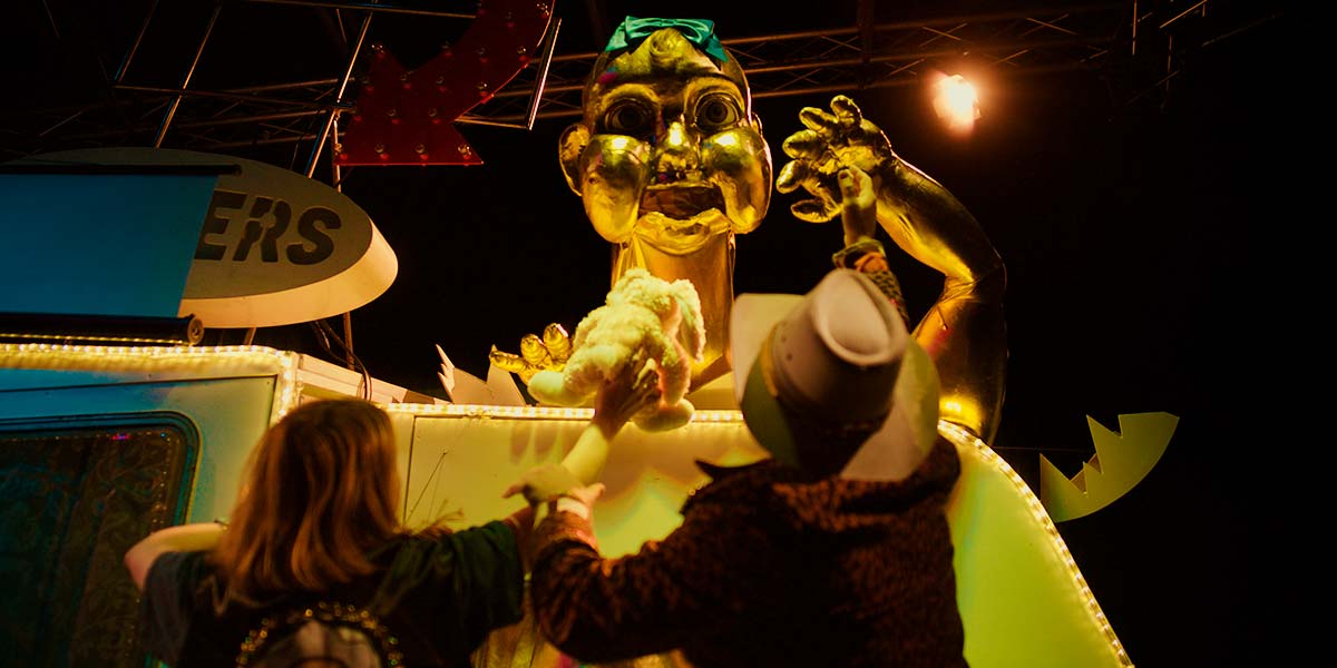 A giant golden baby erupts out of the top of a caravan. It holds one arm in the air whilst two people stand before it, waving a cuddly toy.