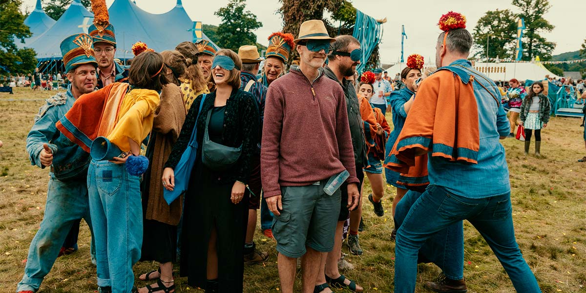 On a festival field, a group of participants clump together, grinning. They wear blindfolds and are surrounded by people dressed in denim with orange capes and large pompoms on their heads.