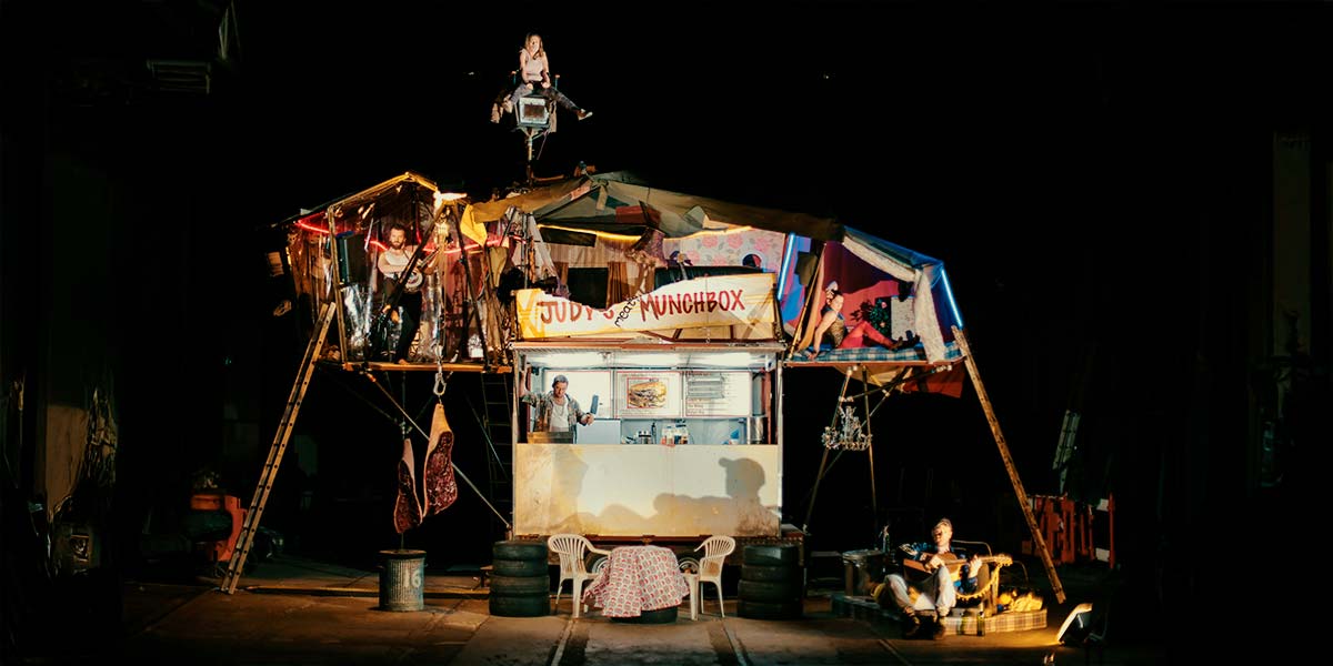A burger van is lit in stark light against a pitch-black sky. It has a ramshackle, tented home perilously perched above it and meat carcasses hanging from hooks at the side. A sign above the van reads ‘Judy’s Meaty Munchbox’. Shady-looking characters lurk around the burger van and rooms above. High above the structure, a young woman sits precariously on a chair attached to a slender pole.