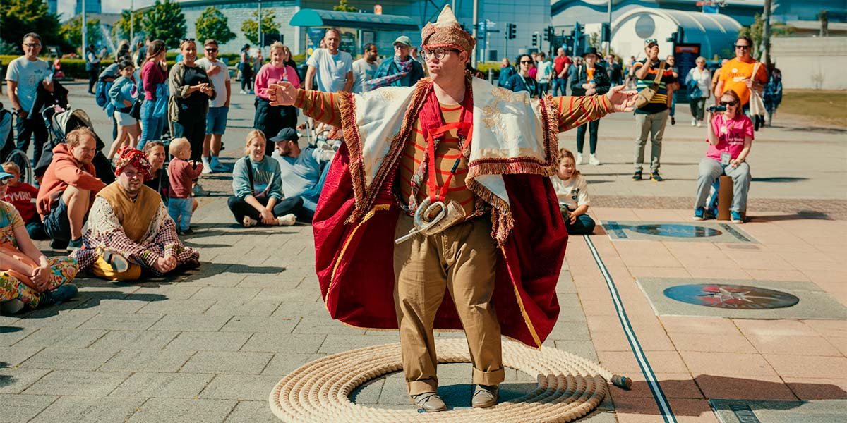 Image description: A man in a red and white billowing cape stands inside a coil of rope. His arms are flung wide and a bugle hangs at his waist. In the background a crowd watch.