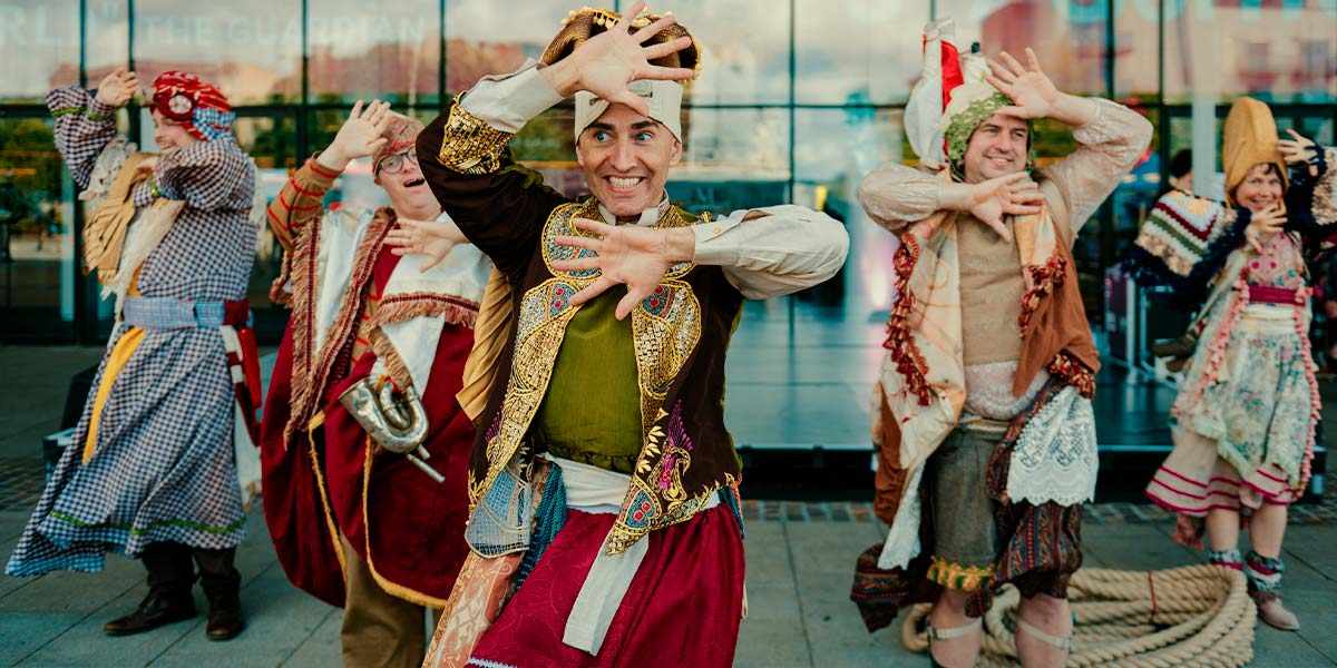 Five performers in colourful, tasselled costumes grin gleefully from behind their splayed fingers.