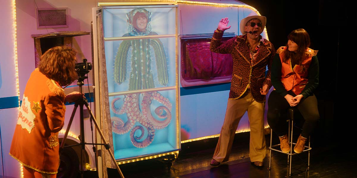 A performer focuses a video camera on an audience member who sits on a stool, whilst another performer talks into a headset. On a screen in the centre is an image of the audience member’s head, imposed onto a cactus body and octopus legs.