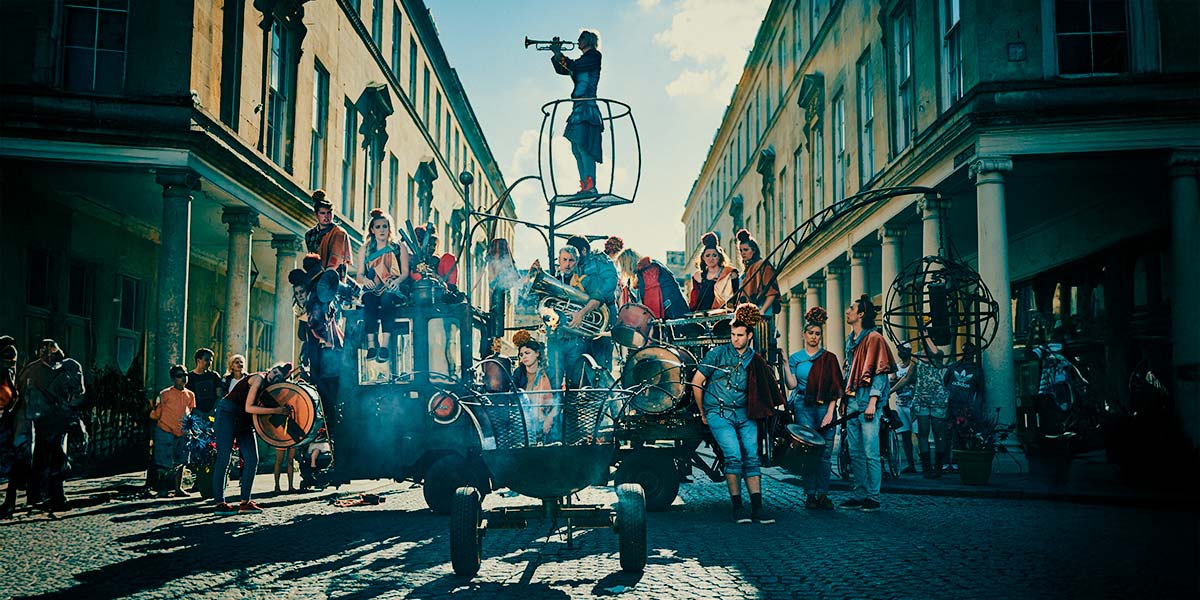  A large group of people dressed in denim with troubadour cloaks over one shoulder and large pompoms on their heads, stand on and around a rust coloured, open-backed vehicle. Some carry brass instruments and drums. From a small crane at the rear dangles a metal globe on a chain and from the centre a raised platform holds someone playing a trumpet. In the foreground, smoke smoulders from a large firepit on wheels.