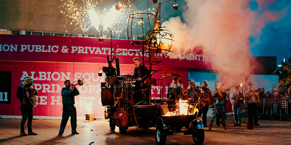 A group play brass instruments around a rust-coloured vehicle. Fire burns from a cage hanging above the vehicle and from a fire pit attached to the back. On the platform body of the vehicle, a man plays drums, whilst fireworks erupt in the background.