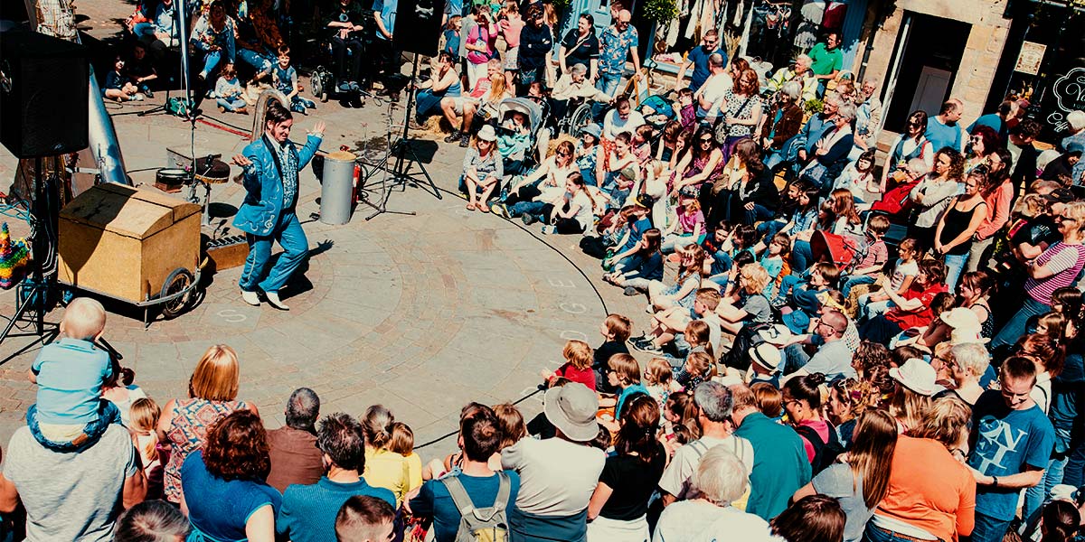 A large audience watch a man in an electric blue suit. He dances inside a circular playing space, in a sunny town square.