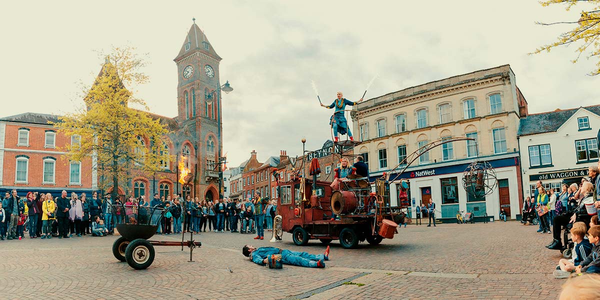 In a town square, a man lies on his back on the ground whilst fire burns from a metal pole nearby. Behind him, a woman stands on an elevated platform, above a rust-coloured vehicle, holding a white-flamed flare in each hand.