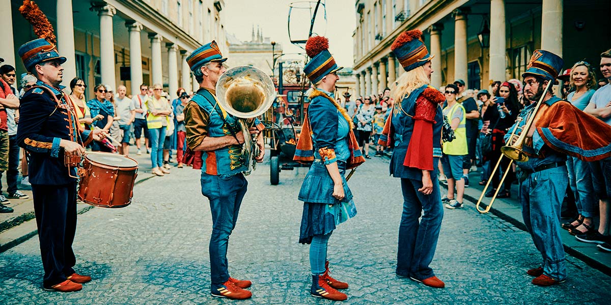 Four band members stand to attention in a line facing another denim-clad band member, who holds a trombone. Behind them, crowds line the pavements.