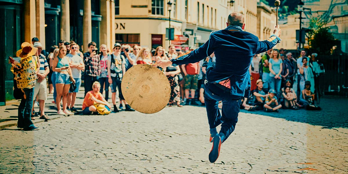 The back of a man dressed in a blue velvet suit leaping into the air, drum stick in one hand, gong in the other, whilst a crowd watches.
