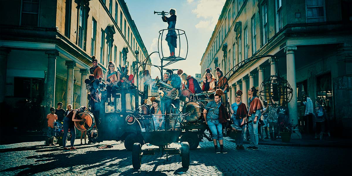 A large group of people dressed in denim with troubadour cloaks over one shoulder and large pompoms on their heads, stand on and around a rust coloured, open-backed vehicle. Some carry brass instruments and drums. From a small crane at the rear dangles a metal globe on a chain and from the centre a raised platform holds someone playing a trumpet. In the foreground, smoke smoulders from a large firepit on wheels.
