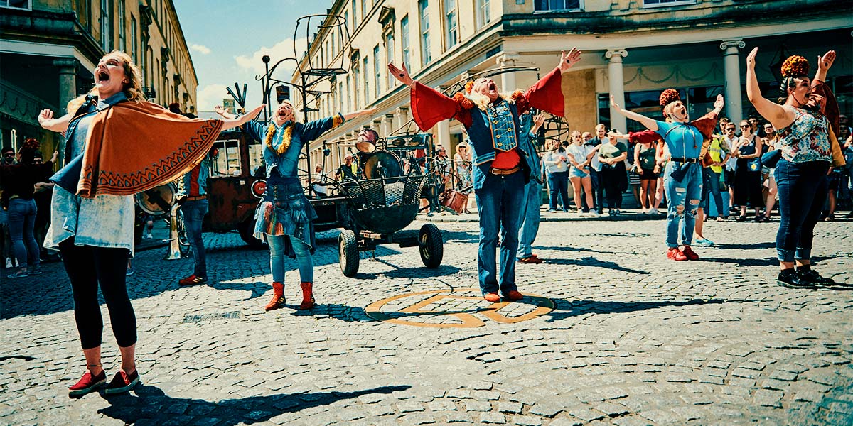 In a town square, scattered performers in various degrees of denim, hold their arms upwards and shout joyfully to the sky.