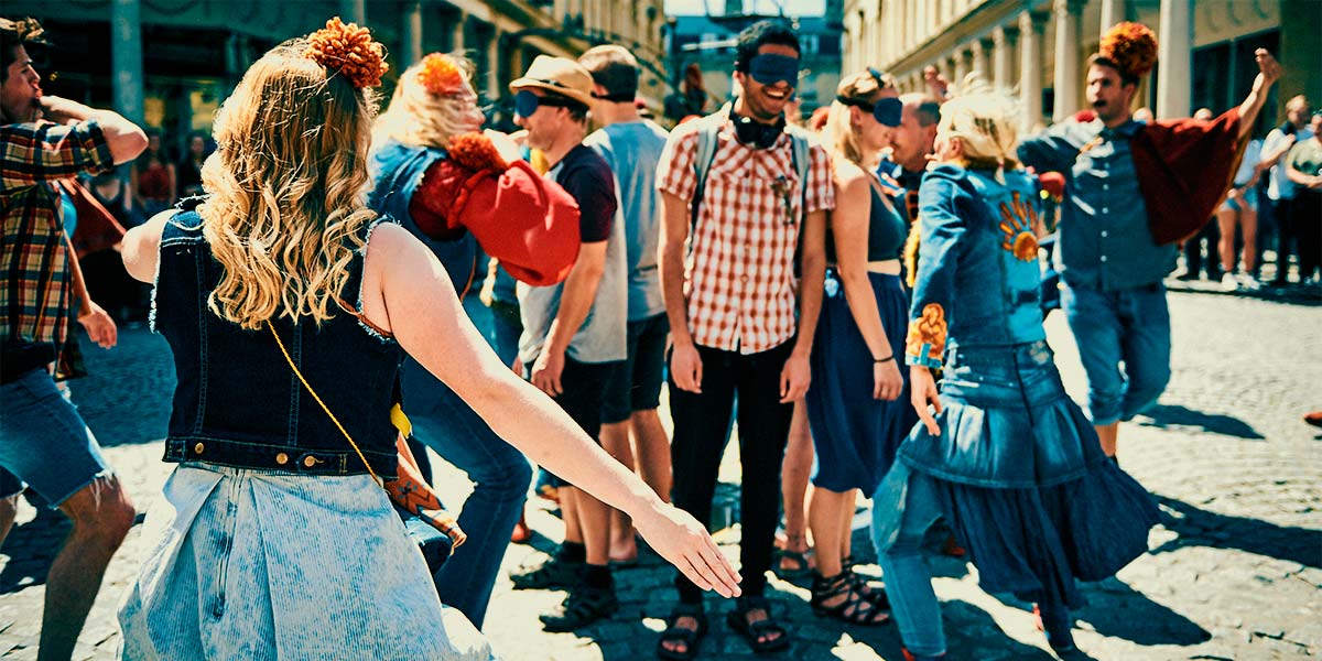 In a town square, a cluster of blindfolded participants are surrounded by dancing performers dressed in denim.