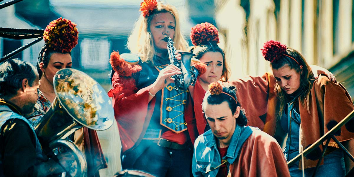 A group wearing red capes and pompoms on their heads stare down with sad expressions, whilst a denim-clad woman with sorrowful eyes plays a soprano saxophone.
