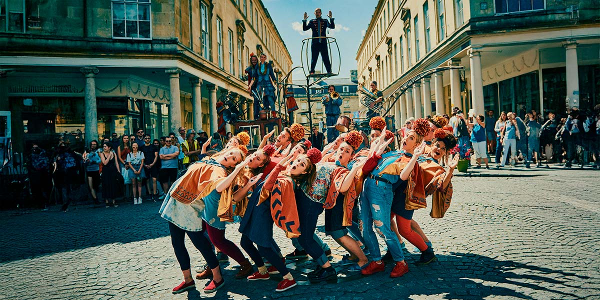  In a town square, a group of young performers lean backwards in a clump, their arms raised at right angles and their mouths open in an ‘o’. They wear orange pompoms on their heads. On an elevated platform behind them, a man in a blue velvet suit has his arms raised in a similar gesture.