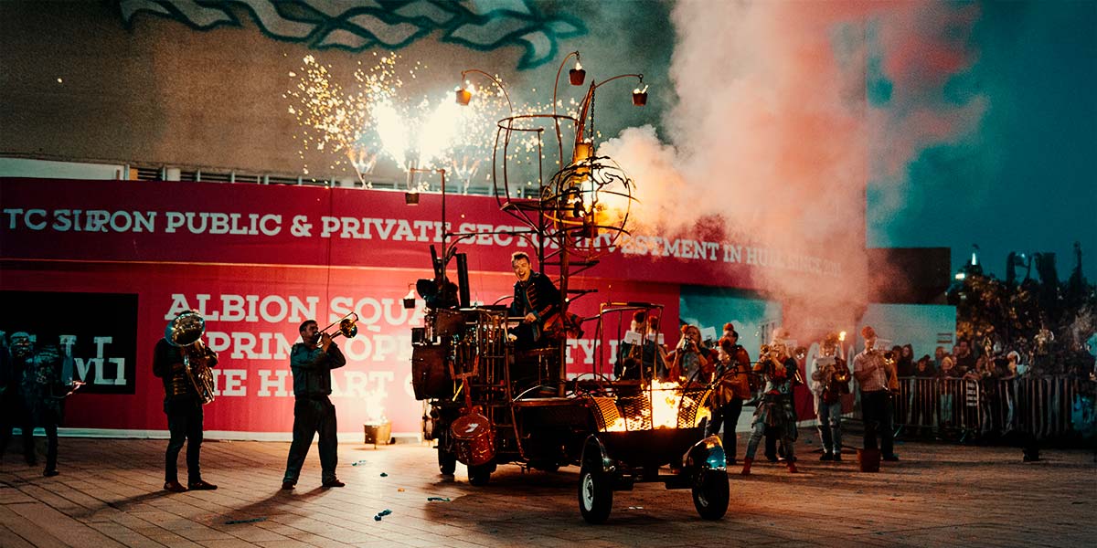 A group play brass instruments around a rust-coloured vehicle. Fire burns from a cage hanging above the vehicle and from a fire pit attached to the back. On the platform body of the vehicle, a man plays drums, whilst fireworks erupt in the background.