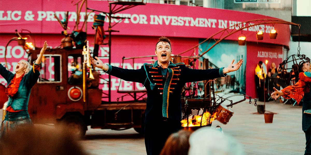 A man in a blue velvet suit stands with his arms outstretched and mouth open as he shouts to the sky. Behind him, a fire pit burns whilst pots of fire hang from a crane attached to a vehicle.