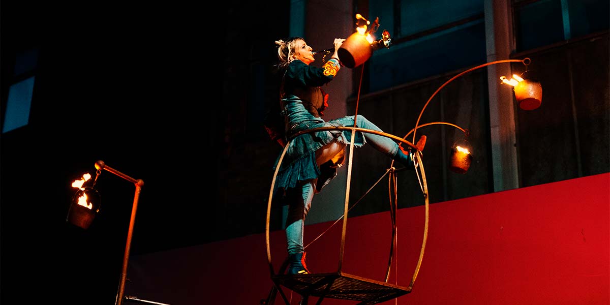 A woman stands on a small platform, one leg resting on the handrail. She plays a trumpet into the night, whilst pots of fire suspended from the platform burn brightly.