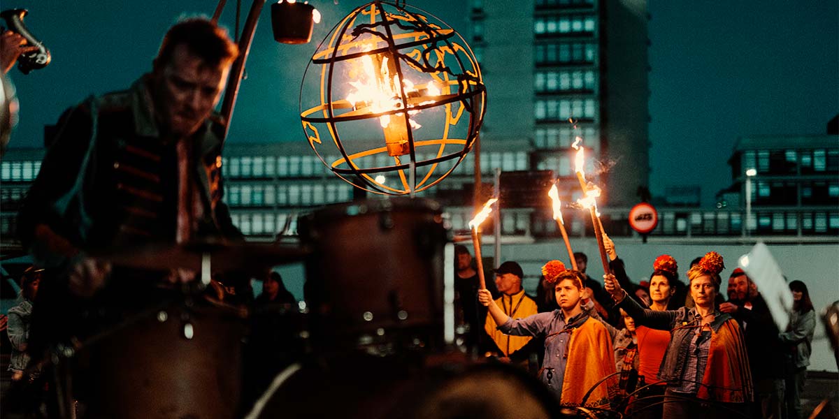 Under a night sky, a group wearing orange capes and pompoms on their heads hold up flaming torches. Their faces are sad in the soft light. In the foreground, a globe-shaped cage holding fire burns brightly, whilst a downcast drummer plays his drums.