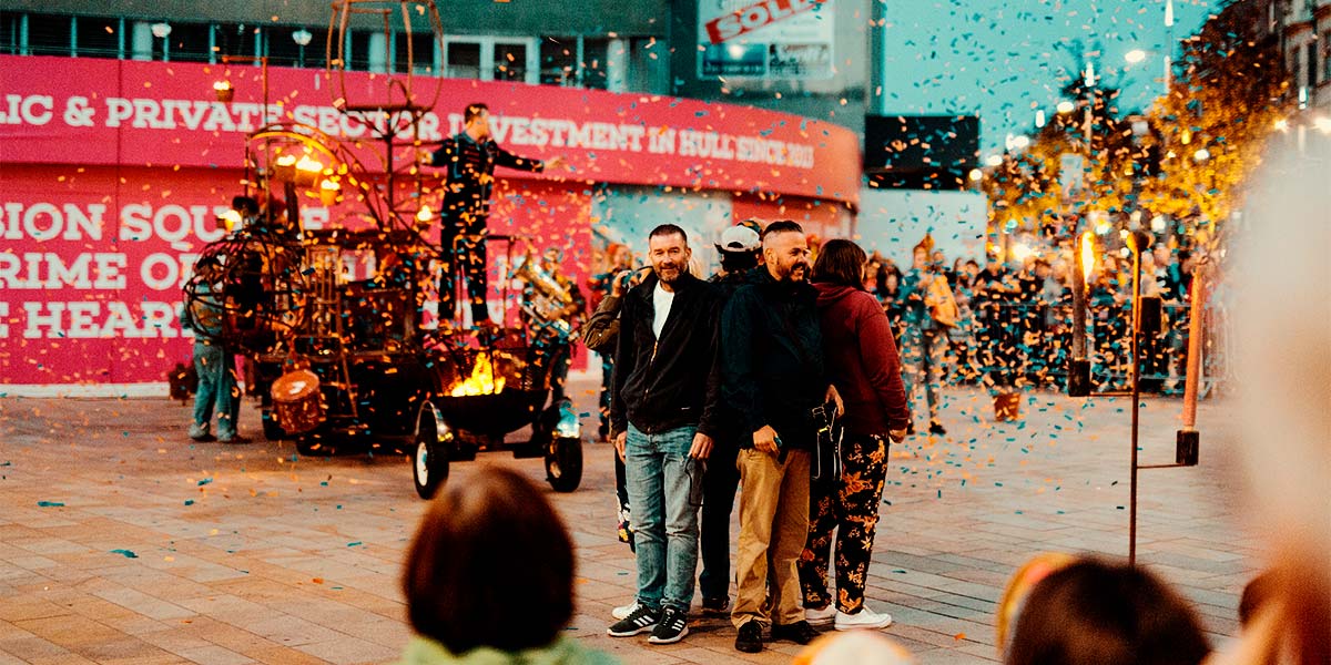 Four people stand back-to-back in a town square, whilst golden confetti flutters around them. In the background a firepit, attached to a vehicle with band members on and around it, burns.