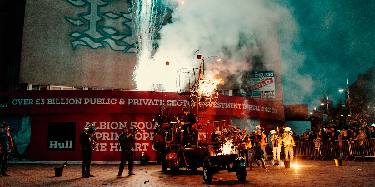 In twilight, a cluster of people play brass instruments around a rust-coloured vehicle.  Above it, a globe-shaped cage hangs, with a fire raging inside. Fire burns from a fire pit on wheels at the back of the vehicle and, from the low roof of a building behind, fireworks explodes.