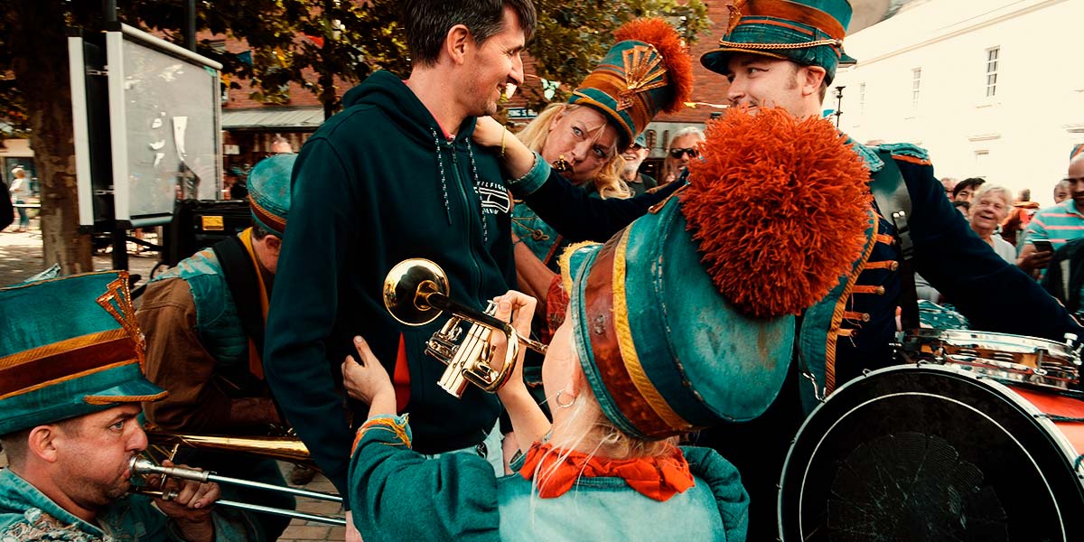 A denim-clad band play their instruments against the body of a smiling participant, whilst the drummer holds their shoulder conspiratorially.