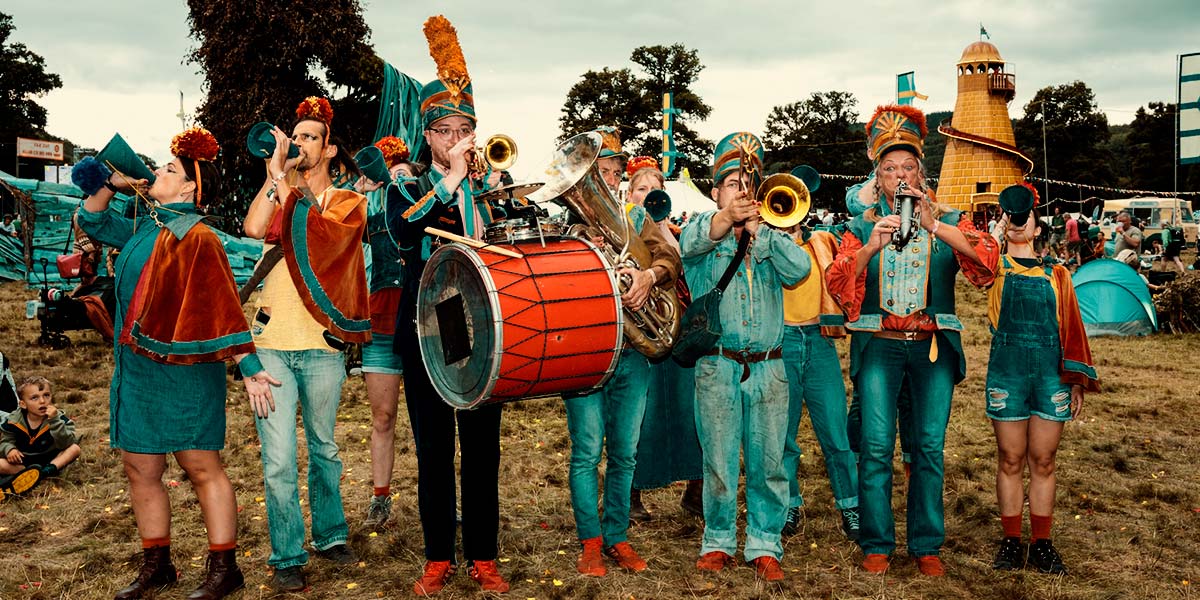 In a festival field, a group of people dressed in denim with red pompoms on their heads, hold brass instruments or blue cones to their mouths.