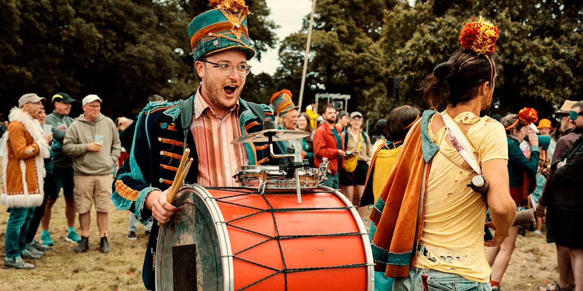 A man with a red drum strapped to his front and wearing a tall denim hat with an orange pompom plume, strides around a field with his mouth open in a gleeful shout. Around him, performers wearing pompoms on their heads move amongst a throng of people.