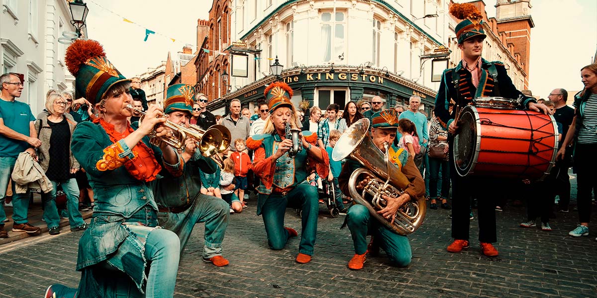 In a town square surrounded by audience, the brass band kneel in a horseshoe and play their instruments, whilst the drummer stands to attention holding a red bass drum.