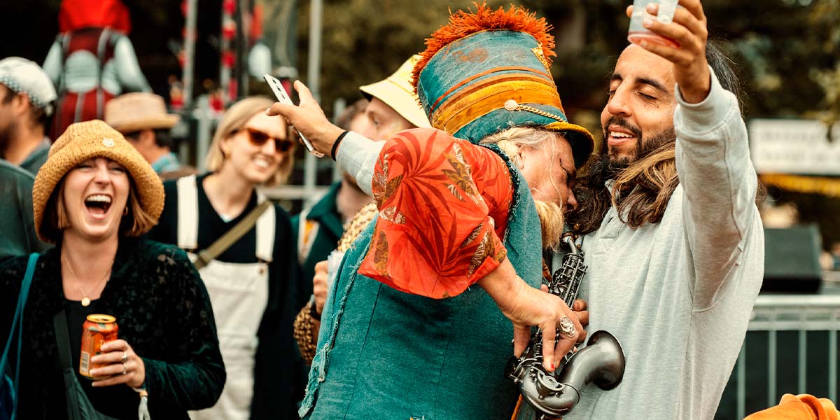At a festival, a woman dressed in denim plays her saxophone against the belly of a participant. They receive it with closed eyes, arms outstretched and an empty pint cup in one hand, whilst onlookers grin.