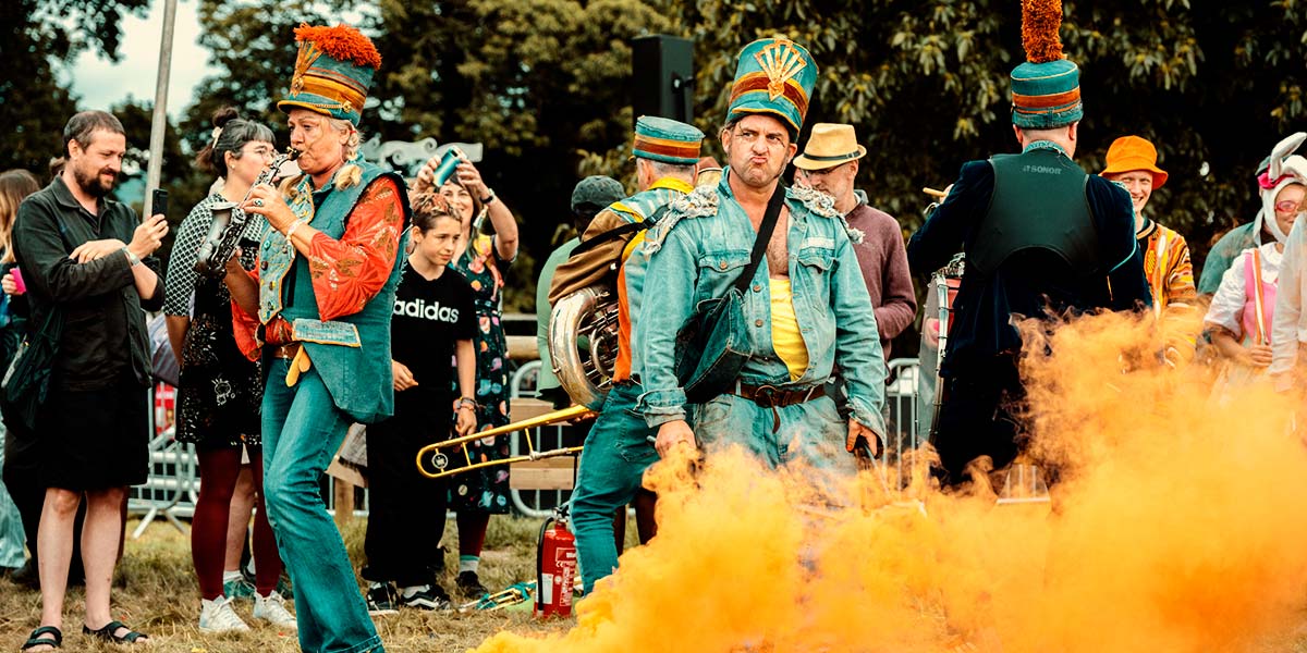 In a field, the band play their instruments to smiling onlookers. One man band member pouts his lips behind a bucket that emits plumes of orange smoke.