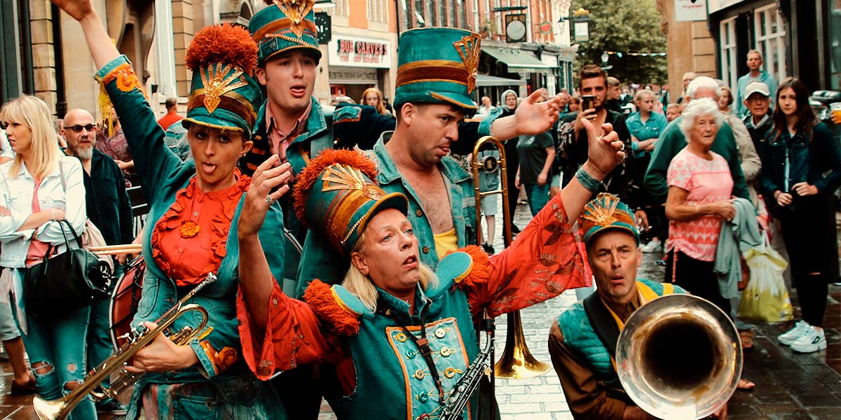 On a street, the band pose with their instruments. They wear double denim accessorised with red, and tall denim hats with a gold emblem and giant pompoms on top. They stick their tongues out and pull dopey expressions.