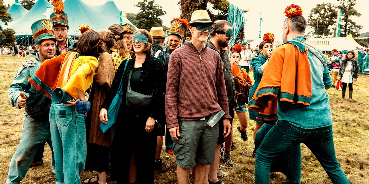 On a festival field, a group of participants clump together, grinning. They wear blindfolds and are surrounded by people dressed in denim with orange capes and large pompoms on their heads.