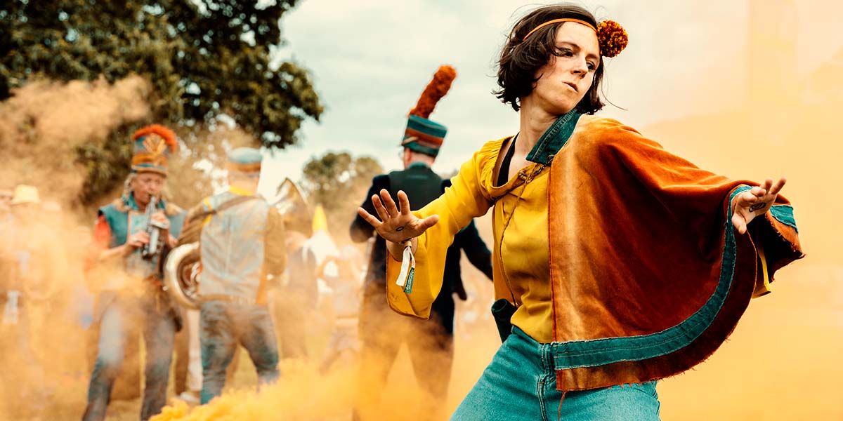 A performer with an orange pompom tied around their forehead, swishes an orange cape fixed at their shoulder.  Behind them, orange smoke erupts from a bucket as musicians play brass instruments.