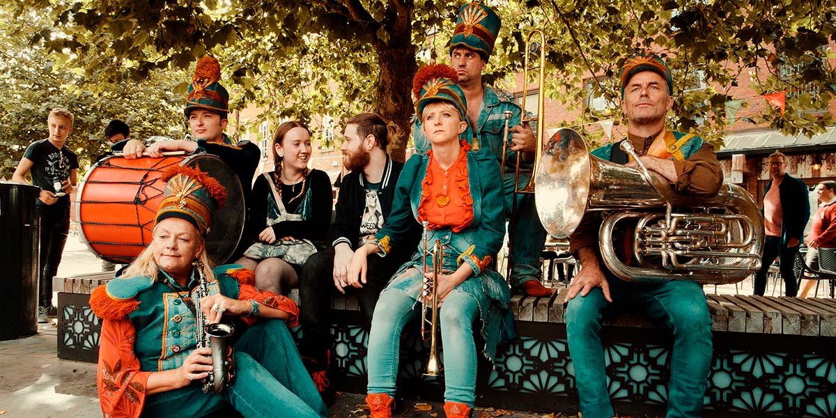  Two people sit on a bench, smiling at each other. Posed around them are five musicians holding brass instruments and a bass drum. They wear double denim, red shoes and tall denim hats with large red pompoms on top.