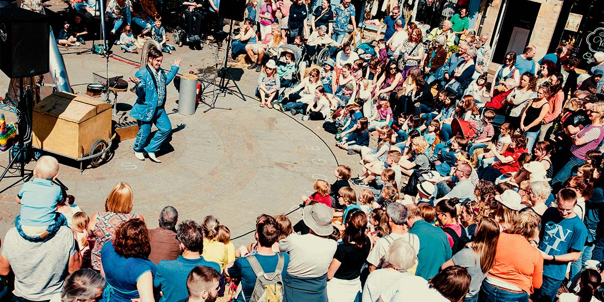 A large audience watch a man in an electric blue suit. He dances inside a circular playing space, in a sunny town square.