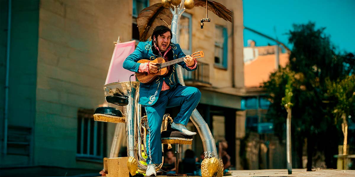 A man in an electric blue suit embellished with a treble clef and white cowboy boots, stands with one foot resting on a stool. He plays a baritone ukulele and sings.