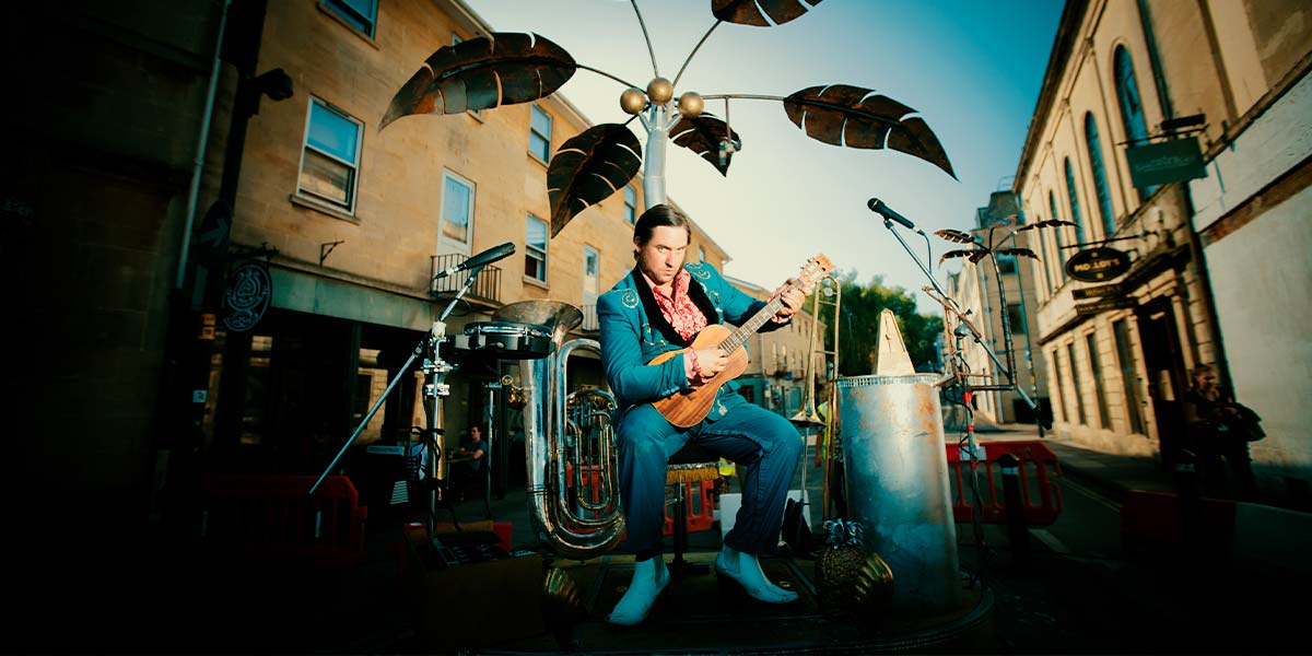 A man with slicked hair and suave electric blue suit holds a bass ukulele and poses on a street. Clustered around him is an assortment of instruments and microphones, whilst above, a metal palm tree opens its fronds to the sky.