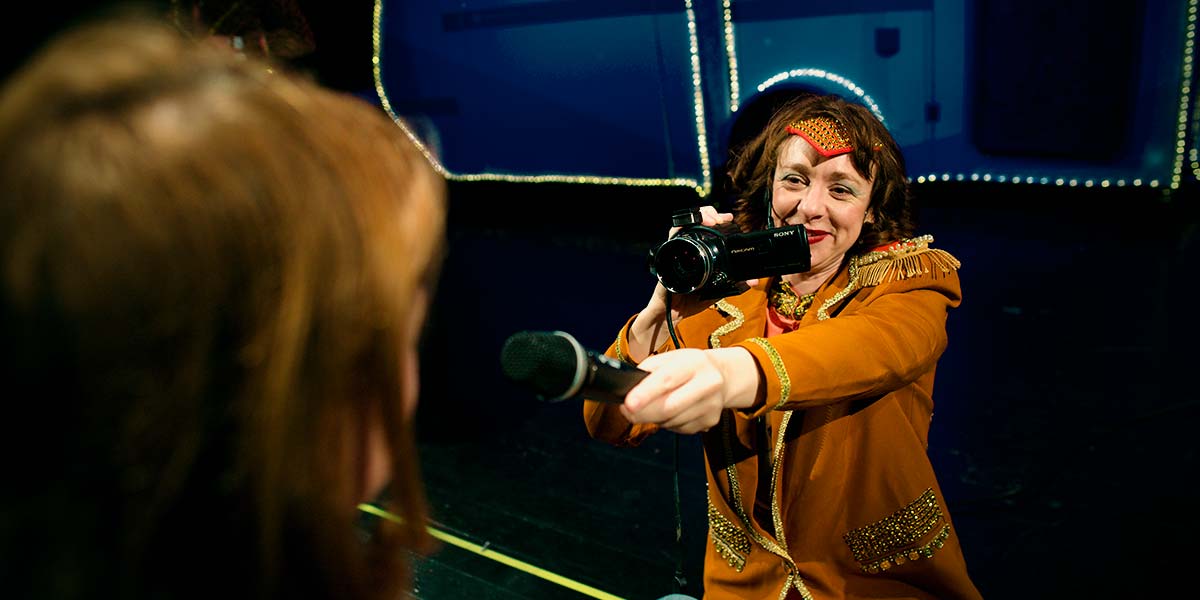 A smiling performer points a microphone towards an audience member, whilst filming with a video camera.