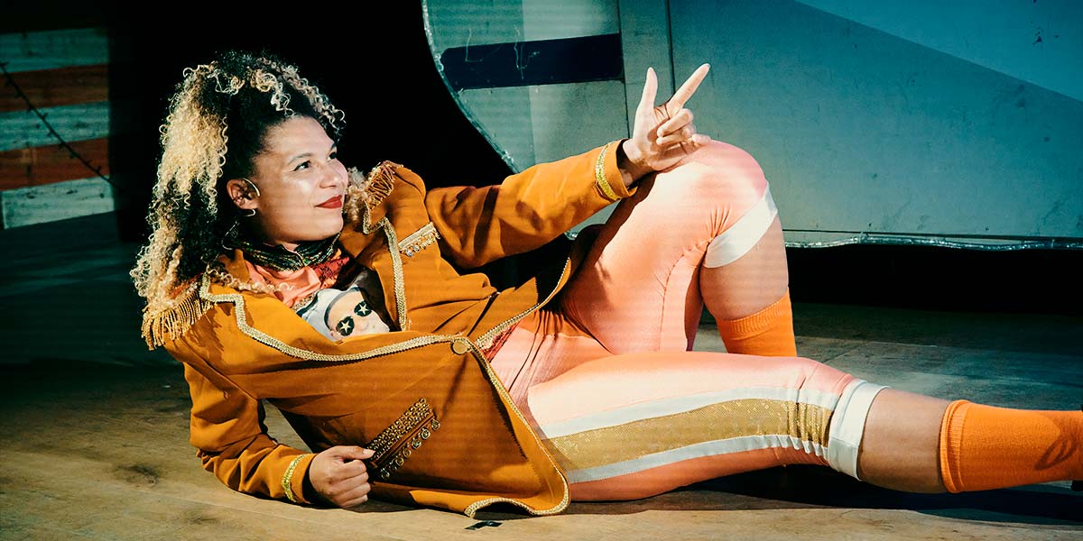 A smiling woman, dressed in an orange blazer and leggings, lies casually on the floor with one knee raised. She rests a hand on top of her bent knee and points with her finger and thumb.