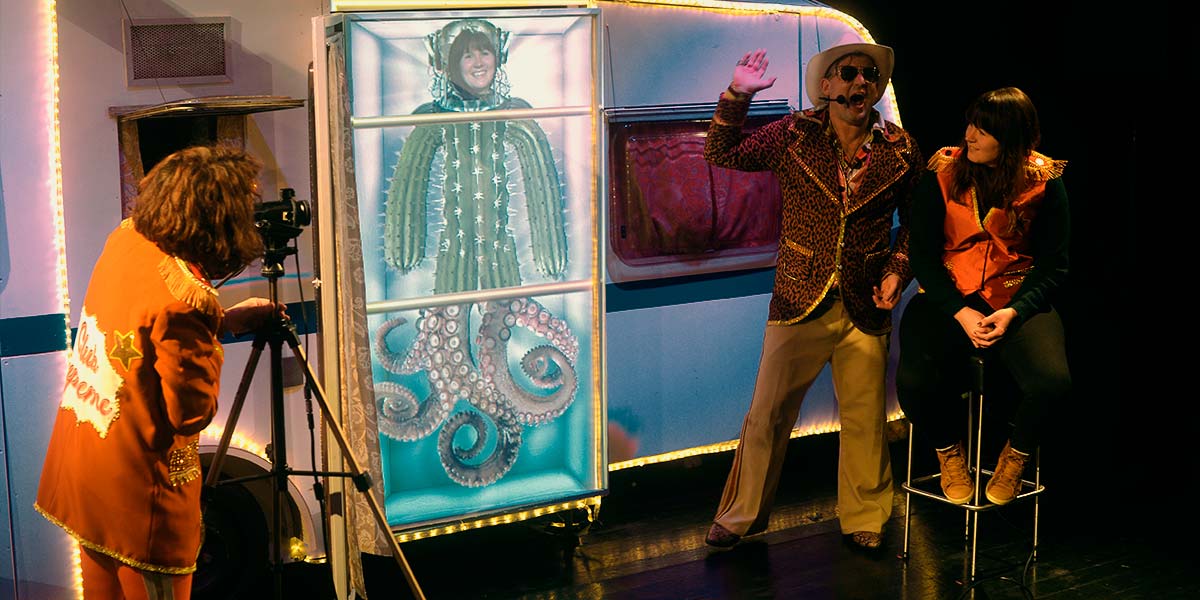 A performer focuses a video camera on an audience member who sits on a stool, whilst another performer talks into a headset. On a screen in the centre is an image of the audience member’s head, imposed onto a cactus body and octopus legs.