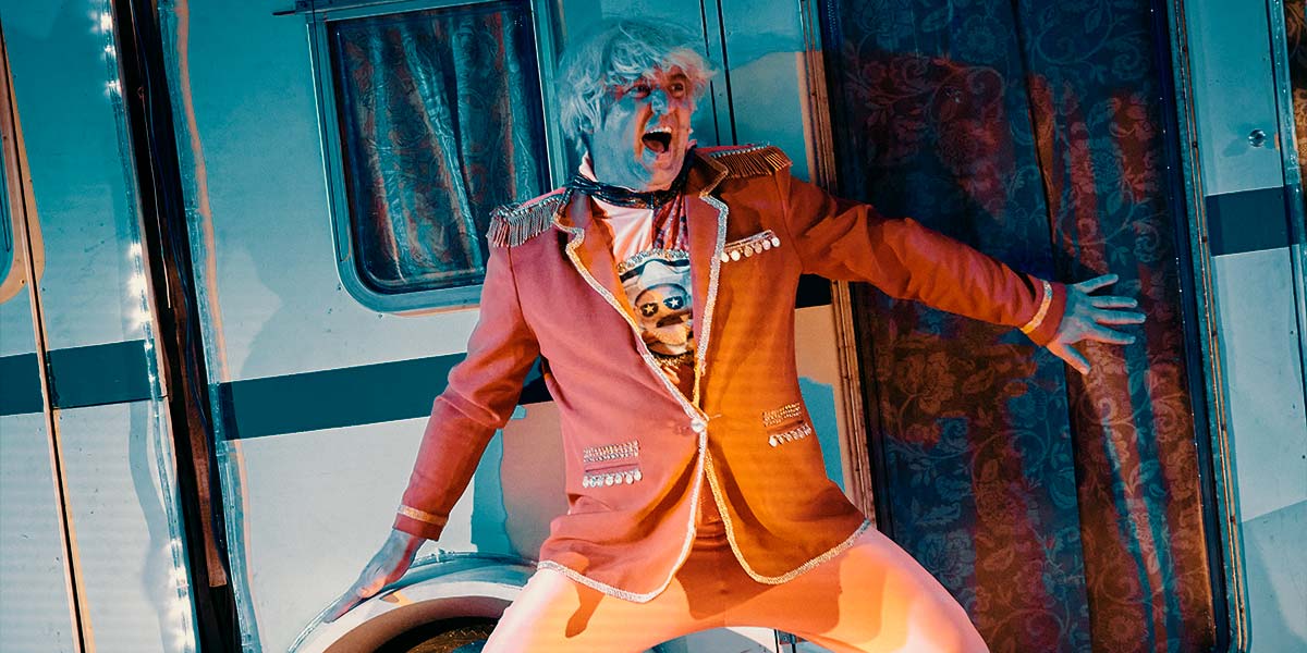  A man in an orange blazer and leggings squats against a caravan; one hand across the door. His mouth is open in an expression of excitement.